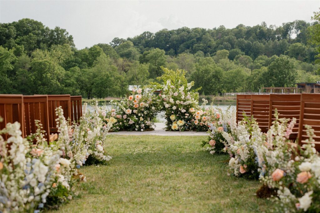 Outdoor ceremony seating space with grassy aisle, floral arrangements, floral arbor. There is a lake and forest trees in background.