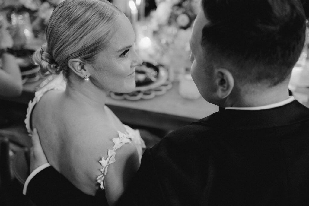 Black and white image. Bride and groom share a glimpse at each other while seated at head reception table.