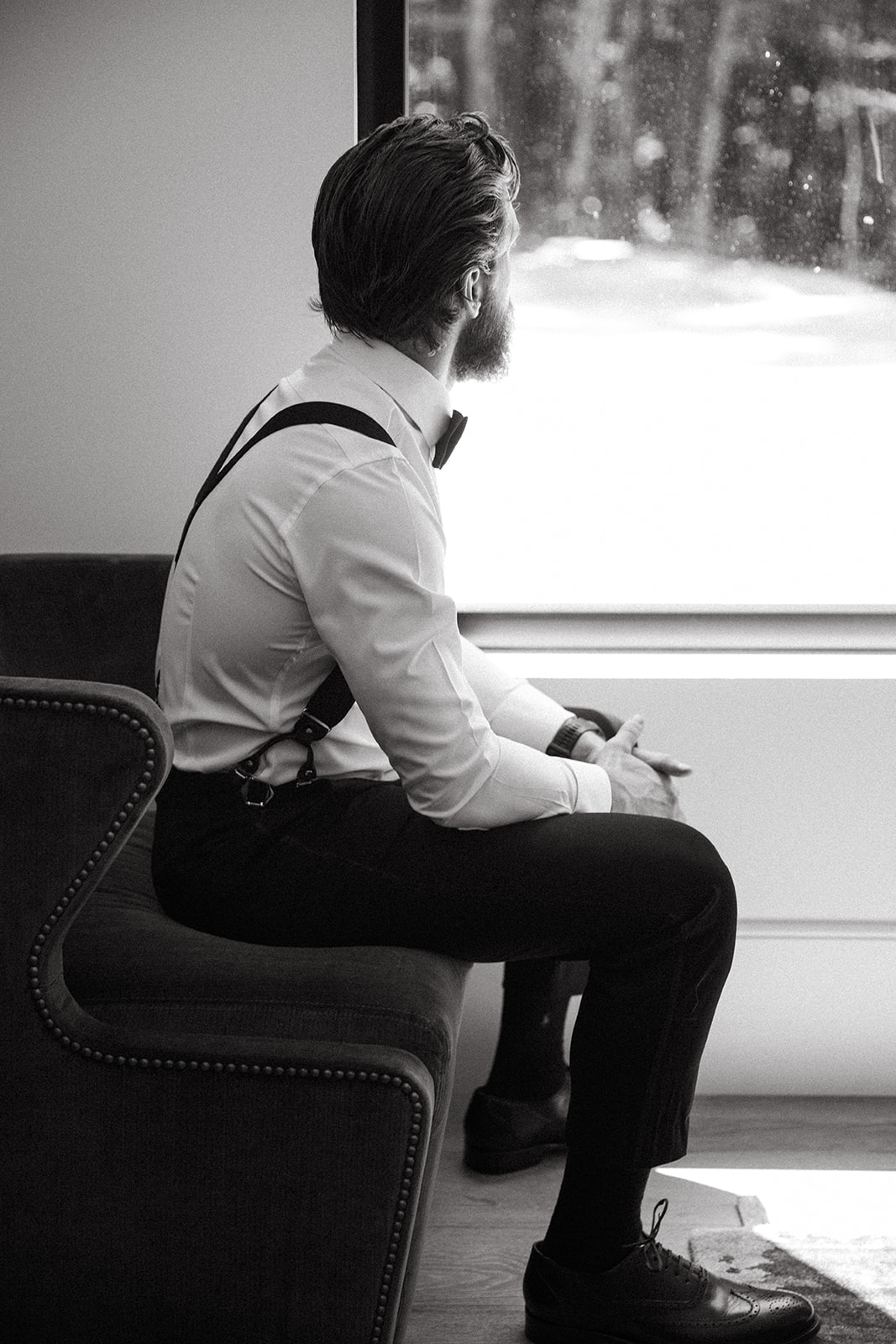 Groom seated on chair looking out window.